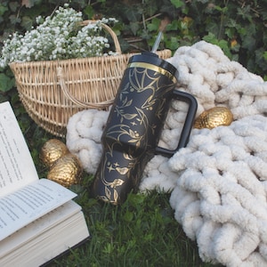 May include: A black and gold stainless steel tumbler with a floral design and a straw. The tumbler is sitting on a white blanket with a book and a basket of flowers.