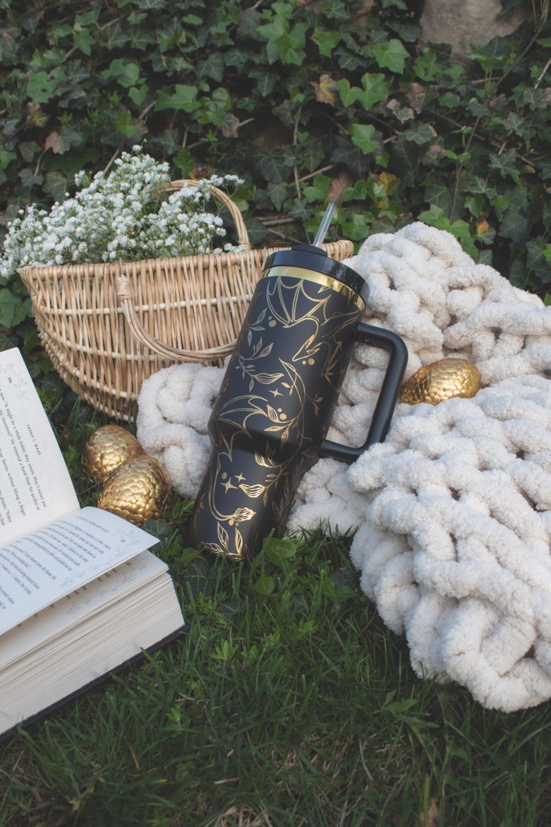 May include: A black and gold stainless steel tumbler with a floral design and a straw. The tumbler is sitting on a white blanket with a book and a basket of flowers.