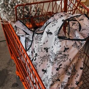 Peut inclure: Un sac de courses r&eacute;utilisable blanc avec un imprim&eacute; noir et blanc de chauves-souris, d'&eacute;toiles et de fleurs. Le sac est dans un chariot rouge.