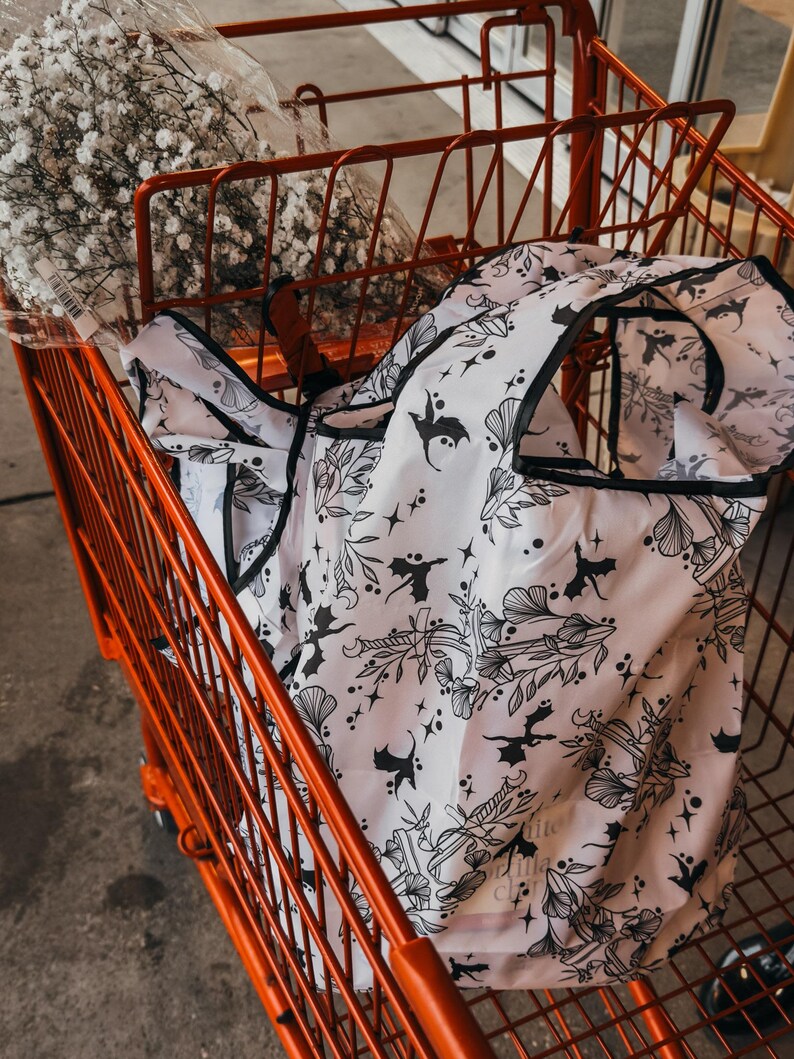 Peut inclure: Un sac de courses r&eacute;utilisable blanc avec un imprim&eacute; noir et blanc de chauves-souris, d'&eacute;toiles et de fleurs. Le sac est dans un chariot rouge.