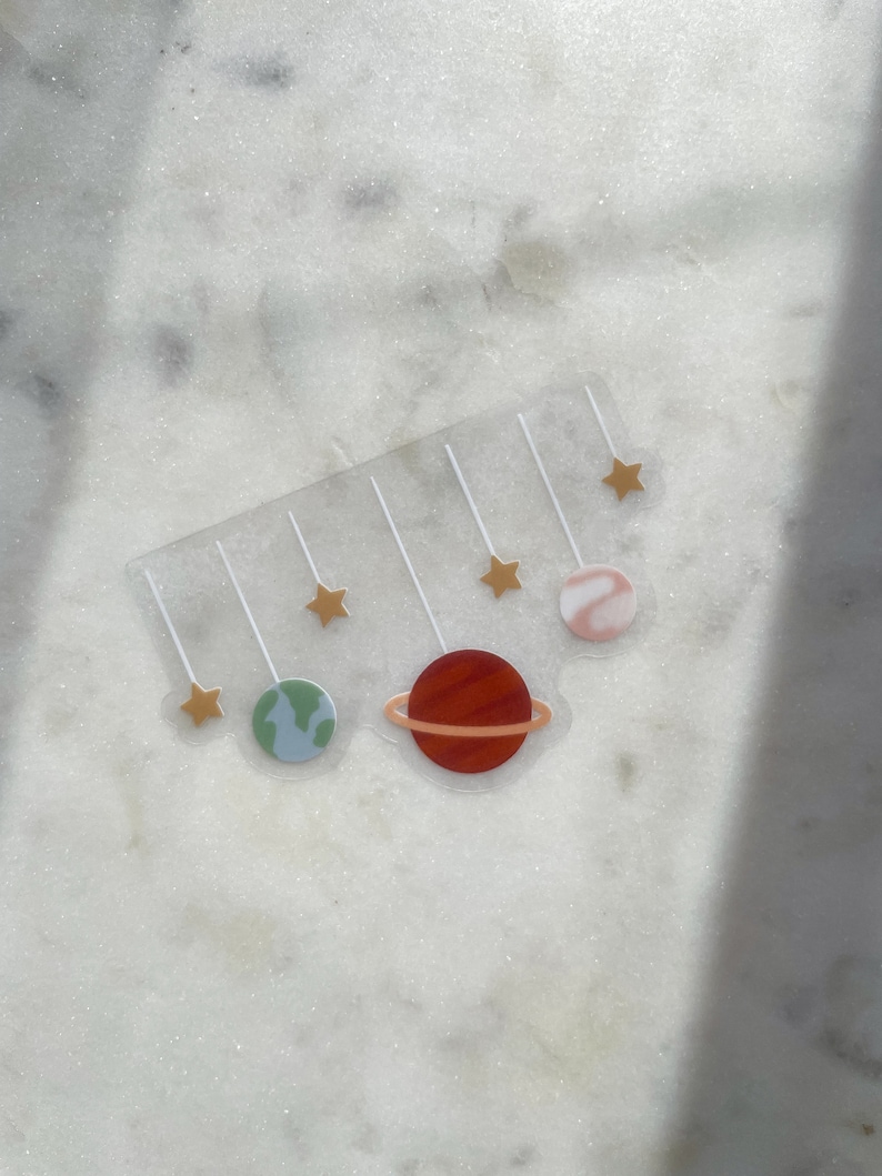Pu&ograve; includere: Un foglio di adesivi trasparente a tema spaziale. Presenta cinque stelle dorate, un pianeta rosso con un anello, un pianeta verde e blu e un pianeta rosa e bianco.
