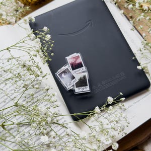 May include: A black e-reader with the Amazon logo rests on an open book, adorned with white flowers. Two small, clear stickers with landscape images are on the e-reader. A golden decorative sphere is in the background.