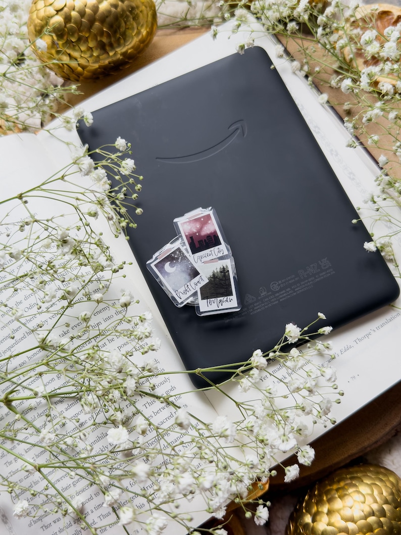 May include: A black e-reader with the Amazon logo rests on an open book, adorned with white flowers. Two small, clear stickers with landscape images are on the e-reader. A golden decorative sphere is in the background.