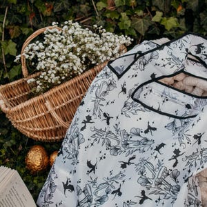 Peut inclure: Un sac de courses r&eacute;utilisable blanc avec un imprim&eacute; noir et blanc de dragons, de fleurs et d'&eacute;toiles. Le sac est pli&eacute; et pos&eacute; sur un lit d'herbe verte avec un livre et un panier en osier rempli de fleurs blanches.