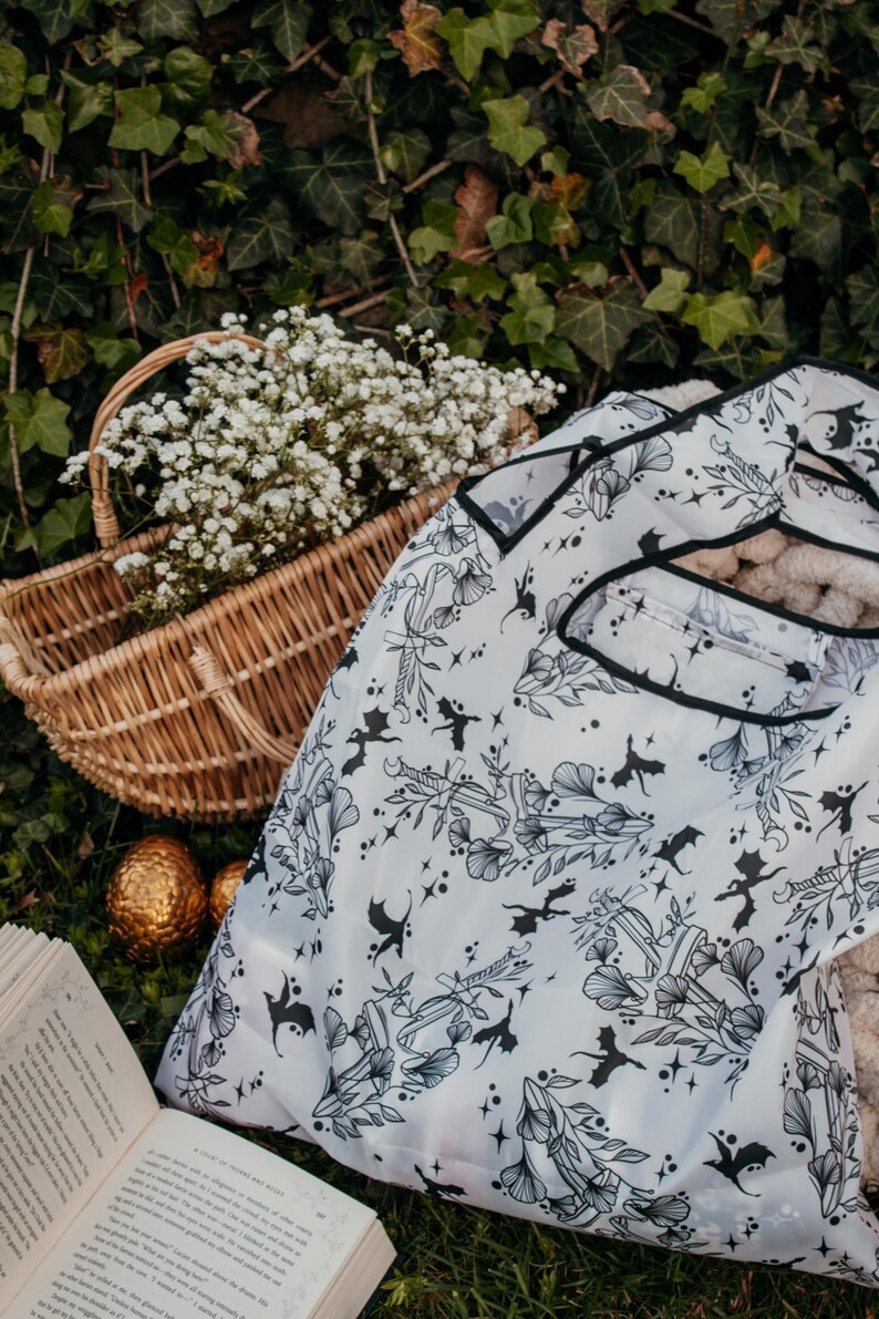 Peut inclure: Un sac de courses r&eacute;utilisable blanc avec un imprim&eacute; noir et blanc de dragons, de fleurs et d'&eacute;toiles. Le sac est pli&eacute; et pos&eacute; sur un lit d'herbe verte avec un livre et un panier en osier rempli de fleurs blanches.