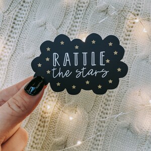 Peut inclure: Un autocollant en forme de nuage noir avec des &eacute;toiles dor&eacute;es et le texte "RATTLE the stars" en blanc.