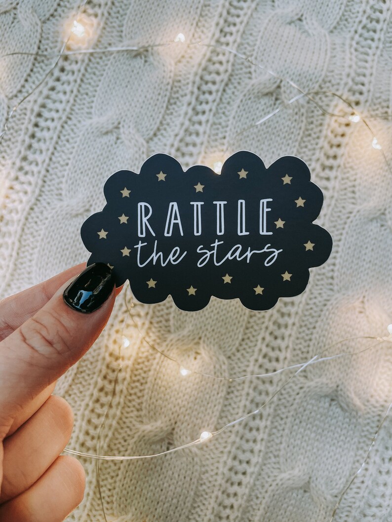 Peut inclure: Un autocollant en forme de nuage noir avec des &eacute;toiles dor&eacute;es et le texte "RATTLE the stars" en blanc.