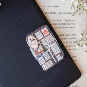 May include: A clear sticker featuring a dragon and various text elements, including "Dying Today Would Be Inconvenient" and "Quest Sword." The sticker is placed on a black e-reader, with a book and floral accents in the background.