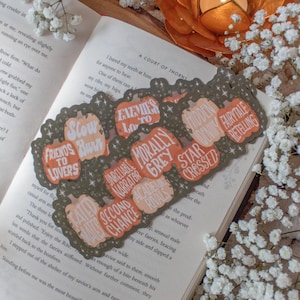 May include: A collection of pumpkin-shaped stickers with phrases like "Friends to Lovers" and "Enemies to Lovers" in orange and white, arranged on an open book. A lit candle sits in a decorative gold holder.