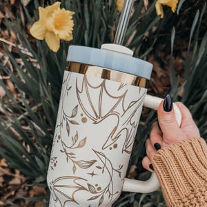 May include: A white stainless steel travel mug with a gold lid and a gold floral design. The mug has a handle and a straw.