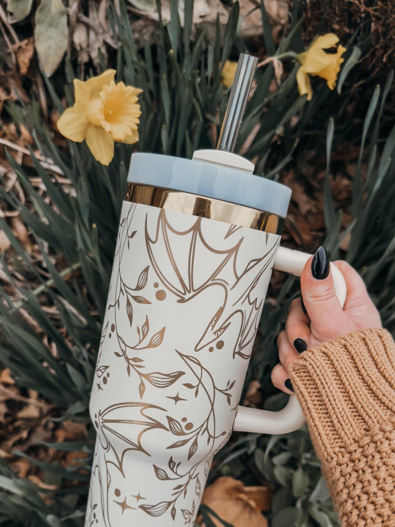 May include: A white stainless steel travel mug with a gold lid and a gold floral design. The mug has a handle and a straw.