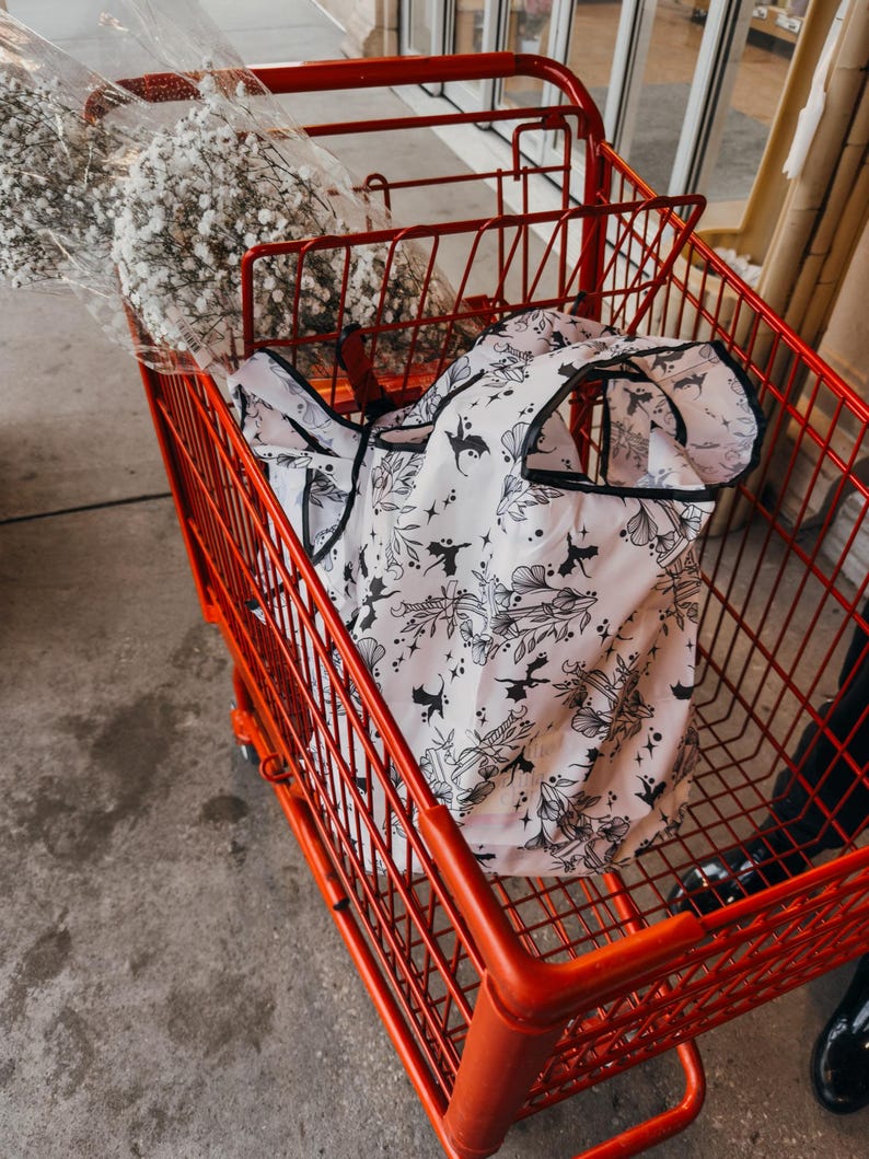 Peut inclure: Un chariot de courses rouge avec un sac de courses r&eacute;utilisable blanc &agrave; motif noir et blanc de dragons et d'&eacute;toiles. Le sac est pli&eacute; et se trouve dans le chariot.