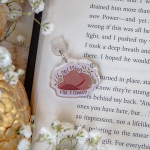 May include: A clear acrylic keychain with a cowboy hat illustration and the text "SAVE A HORSE RIDE A COWBOY." The keychain has a silver-colored clasp and is surrounded by floral elements.