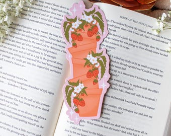 May include: A decorative bookmark showing a stack of terracotta plant pots, each with strawberries and white flowers. The bookmark is set on a book, with a pink border and star details. The book is open.