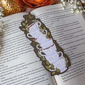 May include: A decorative bookmark featuring a stack of white teacups with the text "Suriel Tea Company" and green leaf accents. The bookmark is set against an open book with text.