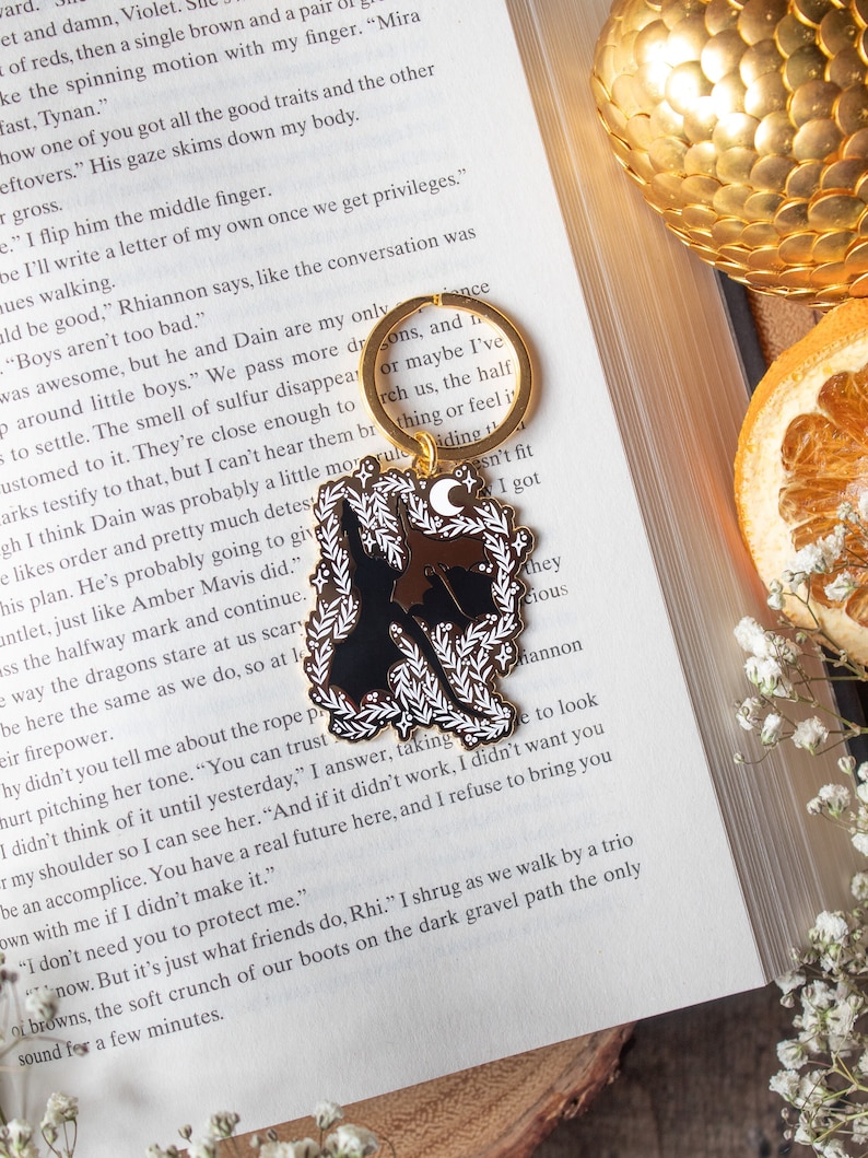 May include: A black and white enamel pin featuring a wolf howling at the moon. The pin is attached to a gold key chain with the words "sons, and if or maybe I've reach us, the half thing or feel it" written on it.