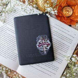May include: A black e-reader with a sticker that reads "you do not yield" is placed on an open book. The scene is decorated with dried orange slices, gold-colored orbs, and white flowers.