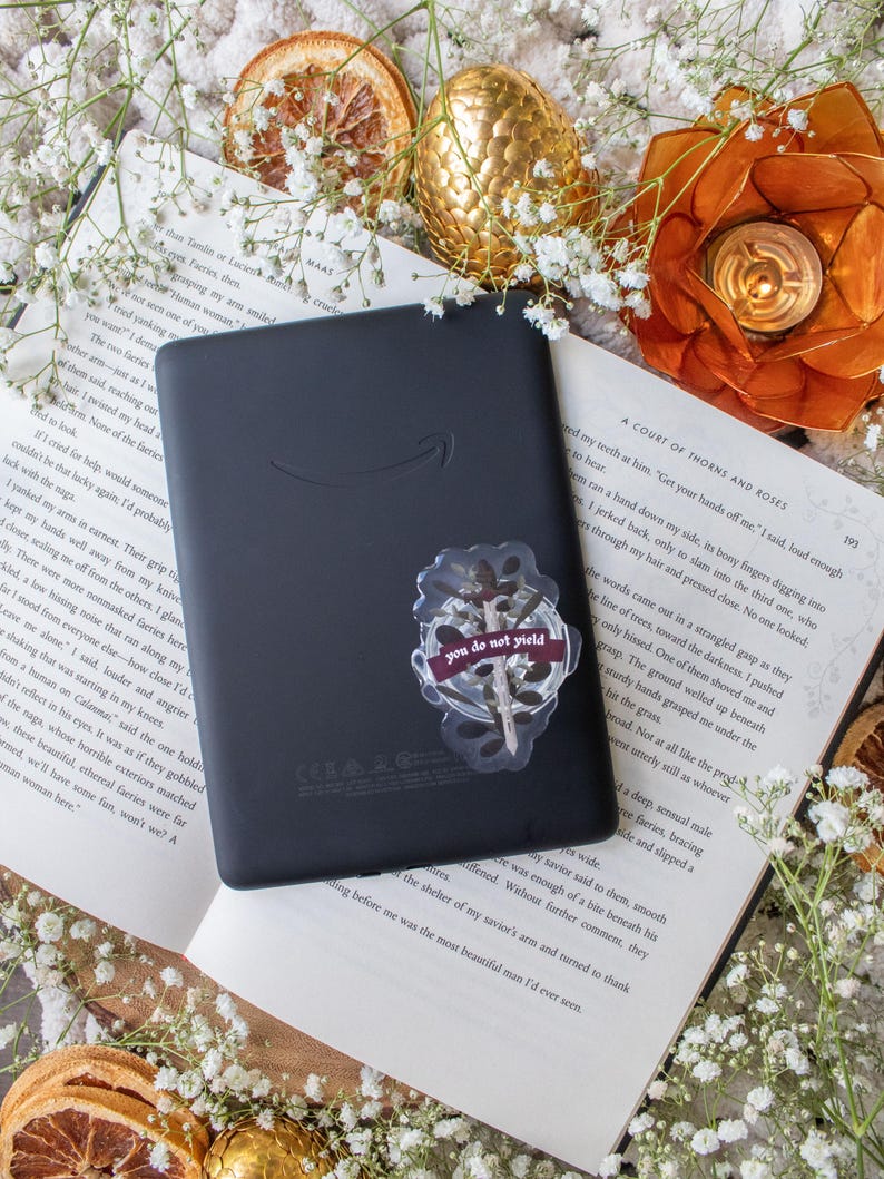 May include: A black e-reader with a sticker that reads "you do not yield" is placed on an open book. The scene is decorated with dried orange slices, gold-colored orbs, and white flowers.