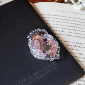 Puede incluir: Un soporte circular transparente para teléfono con una colorida ilustración de una figura con un vestido morado, rodeada de hojas y estrellas naranjas. El soporte está adherido a una tableta negra, apoyada en un libro abierto.