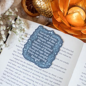 May include: A dark blue bookmark with a quote, on an open book. The bookmark has a decorative border and text that reads "Survival's funny...Keeps me moving forward." Gold and orange floral decorations are in the background.