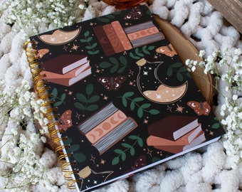 May include: A black spiral bound journal with a dark green, brown, and gold pattern of books, moons, stars, and butterflies. The journal is lying on a white fluffy blanket with a small orange candle holder in the background.