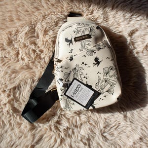 Peut inclure: Sac bandoulière crème avec un motif floral et de chats noirs. Le sac a une sangle noire et une étiquette qui dit "E-READER tech bag". Le sac est sur une surface moelleuse et brun clair.