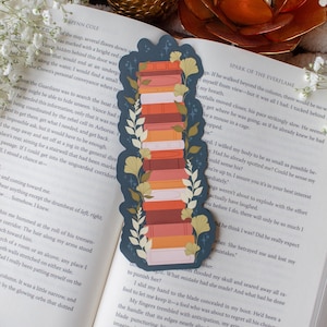 Puede incluir: Un marcapáginas azul marino con una pila de libros en tonos naranja, rojo y rosa. El marcapáginas está decorado con hojas verdes y amarillas y está colocado en un libro.