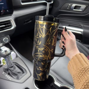 May include: A black and gold travel mug with a floral design and a black handle. The mug is in a car cup holder.
