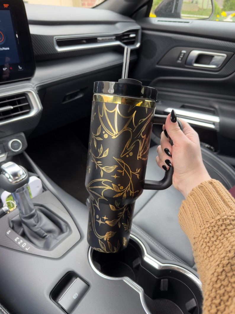 May include: A black and gold travel mug with a floral design and a black handle. The mug is in a car cup holder.