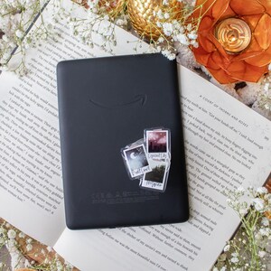 May include: A black e-reader with a sticker set featuring landscape and text designs, resting on an open book. The scene is decorated with dried orange slices, white flowers, and a gold-colored decorative egg.