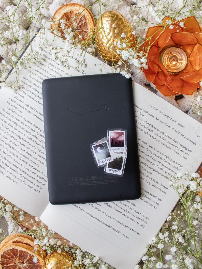 May include: A black e-reader with a sticker set featuring landscape and text designs, resting on an open book. The scene is decorated with dried orange slices, white flowers, and a gold-colored decorative egg.