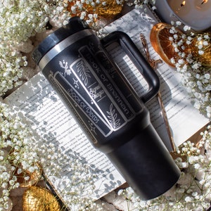 May include: A black stainless steel travel mug with a white design featuring the text "Book of Brennan", "Signet Magic", and "Book of Riders". The mug is sitting on a book with white flowers and a gold dragon egg.