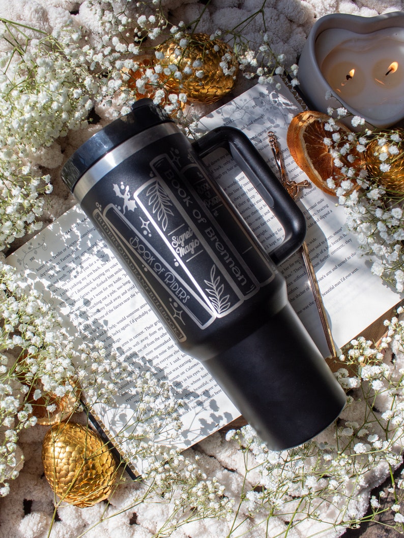 May include: A black stainless steel travel mug with a white design featuring the text "Book of Brennan", "Signet Magic", and "Book of Riders". The mug is sitting on a book with white flowers and a gold dragon egg.