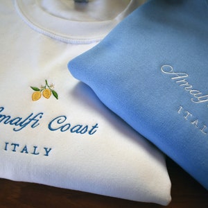 May include: A white sweatshirt and a light blue sweatshirt, each featuring embroidered yellow lemons with green leaves and white blossoms. The text "Amalfi Coast" is embroidered in a cursive script above the block-lettered word "ITALY".