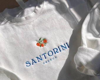 May include: A white crewneck sweatshirt featuring an embroidered design of three orange citrus fruits with green leaves. Below the fruit, the text SANTORINI is embroidered in blue capital letters, with the word GREECE in smaller blue letters underneath.