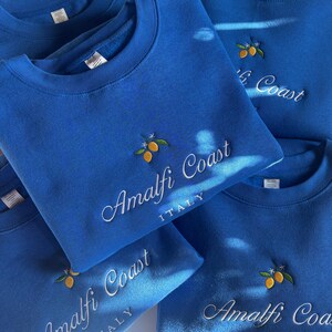 May include: A collection of bright blue crewneck sweatshirts featuring embroidered yellow lemons with green leaves and white blossoms. The text Amalfi Coast is stitched in white cursive script, with the word ITALY in white block letters below.