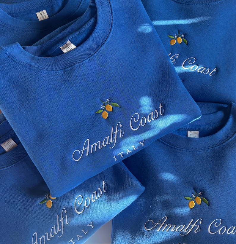 May include: A collection of bright blue crewneck sweatshirts featuring embroidered yellow lemons with green leaves and white blossoms. The text Amalfi Coast is stitched in white cursive script, with the word ITALY in white block letters below.