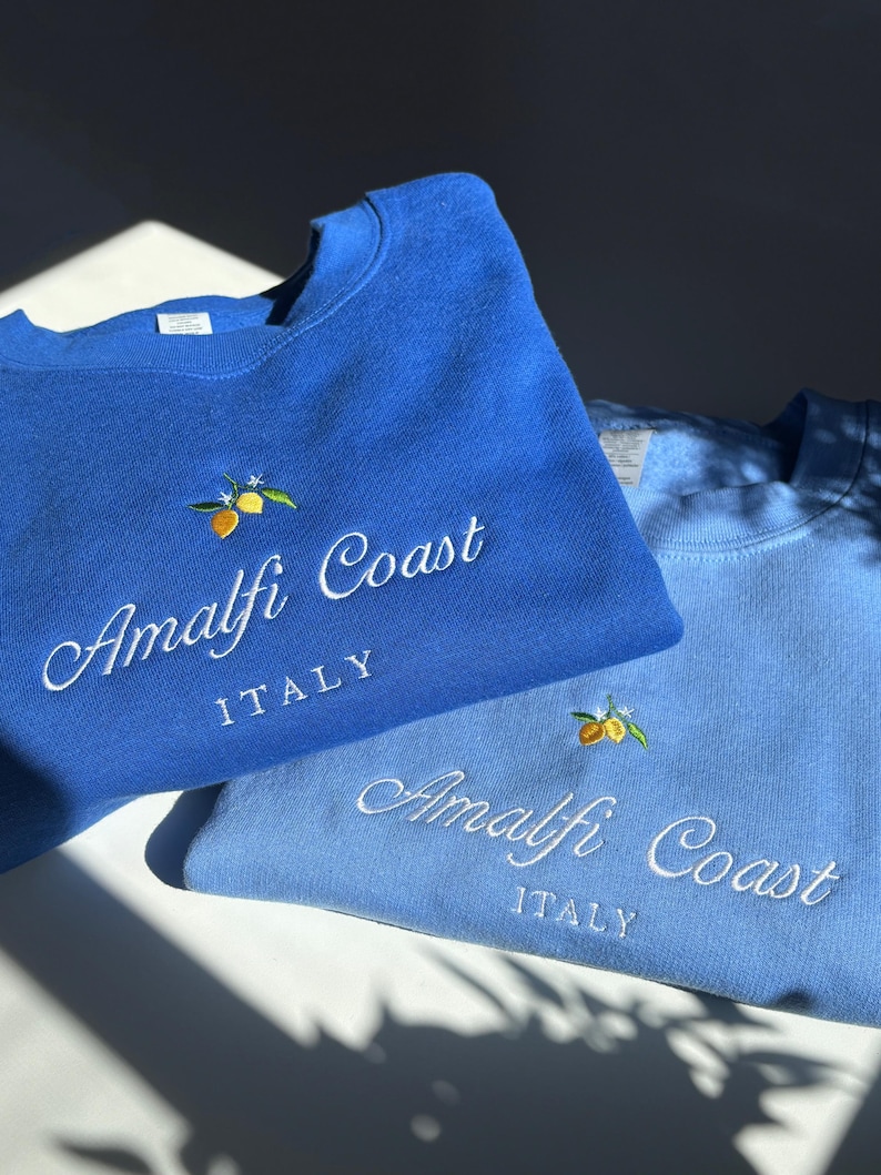 May include: Two crewneck sweatshirts in royal blue and light blue, each featuring embroidered yellow lemons with green leaves and white blossoms above the white cursive text "Amalfi Coast" and the block-lettered word "ITALY" centered on the chest.