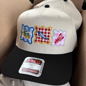 May include: Cream-colored baseball caps with black visors. Each cap features embroidered patches with food-themed designs: lemons, a fish, and a lobster. The caps are in a cardboard box. The tag reads "OTTO CLASSIC FIT OSFM MID PROFILE MAXIMUM COMFORT".
