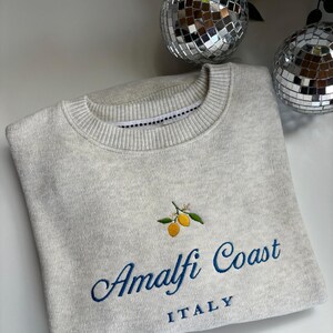 May include: A light gray crewneck sweatshirt featuring an embroidered design of two yellow lemons with green leaves. Below the lemons, the text reads Amalfi Coast in a blue cursive script, with the word ITALY printed in blue block letters underneath.