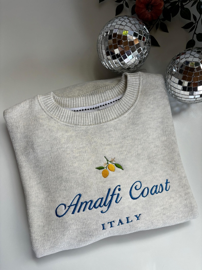 May include: A light gray crewneck sweatshirt featuring an embroidered design of two yellow lemons with green leaves. Below the lemons, the text reads Amalfi Coast in a blue cursive script, with the word ITALY printed in blue block letters underneath.