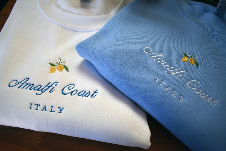 May include: A white sweatshirt and a light blue sweatshirt, each featuring embroidered yellow lemons with green leaves and white blossoms. The text "Amalfi Coast" is embroidered in a cursive script above the block-lettered word "ITALY".