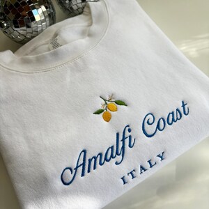 May include: A white crewneck sweatshirt featuring an embroidered design of two yellow lemons with green leaves and small white flowers. Below the lemons, the text reads Amalfi Coast in blue cursive script, with the word ITALY in blue block letters.
