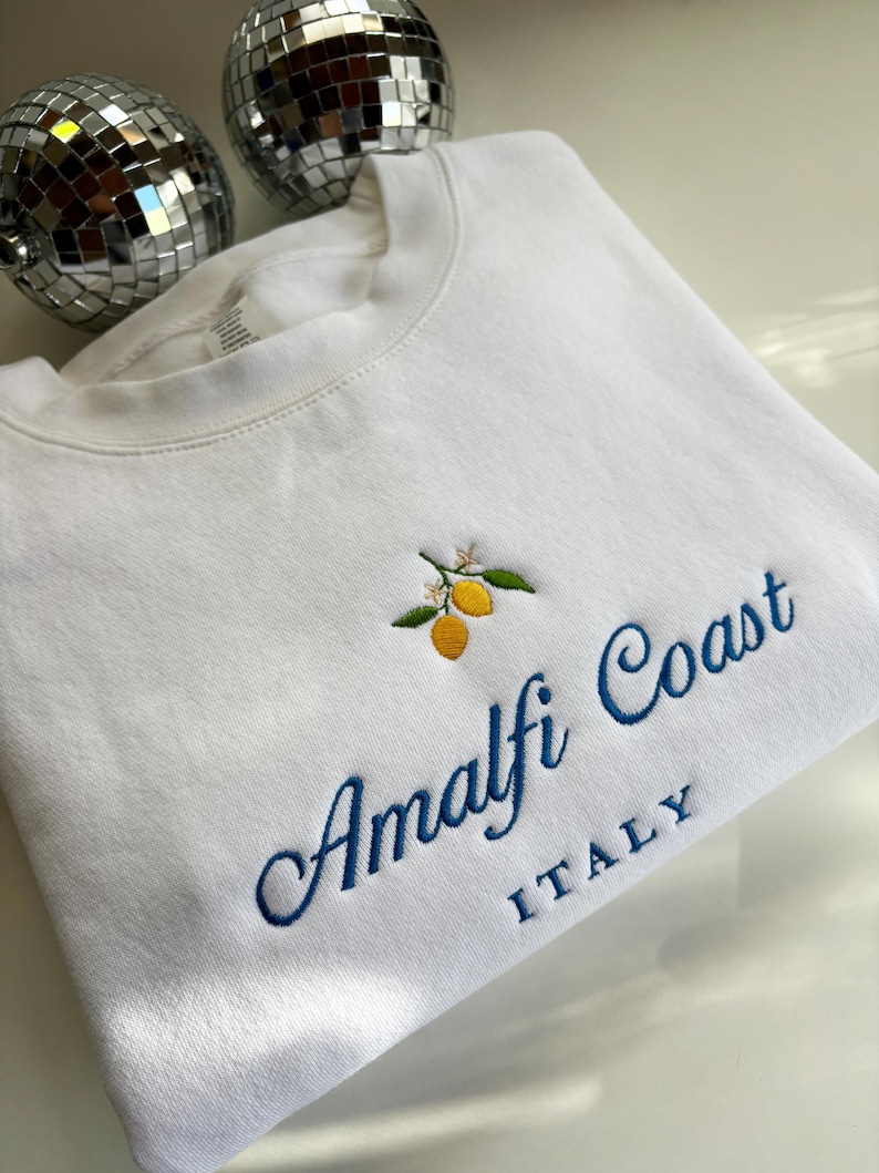 May include: A white crewneck sweatshirt featuring an embroidered design of two yellow lemons with green leaves and small white flowers. Below the lemons, the text reads Amalfi Coast in blue cursive script, with the word ITALY in blue block letters.