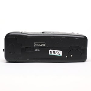 Pode incluir: C&acirc;mara compacta preta com um bot&atilde;o prateado e um adesivo que diz "0952". A c&acirc;mera tem uma etiqueta que diz "Made in China".