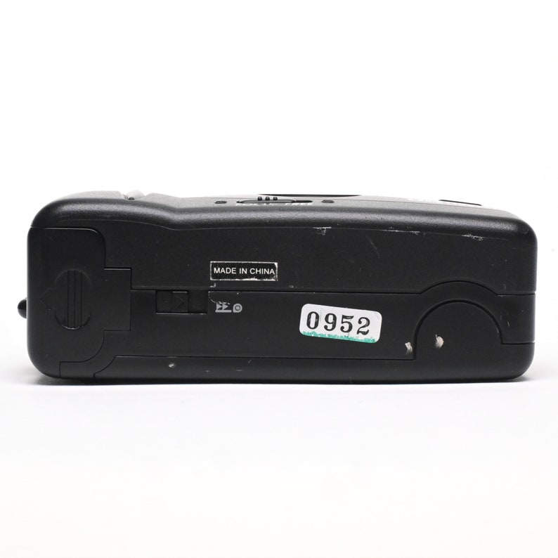 Pode incluir: C&acirc;mara compacta preta com um bot&atilde;o prateado e um adesivo que diz "0952". A c&acirc;mera tem uma etiqueta que diz "Made in China".