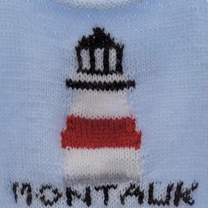 Op de afbeelding: Lichtblau gebreide trui met een vuurtorenontwerp in rood, wit en zwart. Het woord "MONTAUK" is eronder in het zwart geborduurd. De trui heeft een getextureerd uiterlijk.