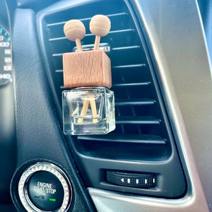 May include: A car air freshener with a clear glass bottle and a wooden top. Two wooden ball-shaped diffusers are inserted into the wooden top. The air freshener is placed in a car's air vent.