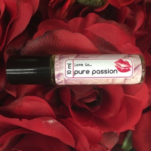 May include: A 10 ml roll-on bottle with a pink and white label that says "Love is... pure passion". The label also features a red lip print. The bottle is sitting on a bed of red rose petals.