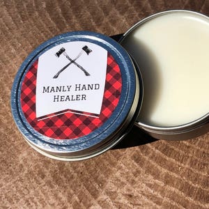 May include: A small tin container with a red and black checked label. The label features a pair of crossed axes and the text "MANLY HAND HEALER". The container is open, revealing a white cream inside.